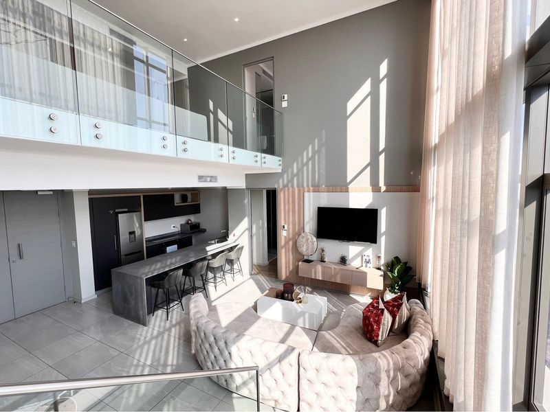 Modern double-volume open-plan penthouse living area featuring a sleek kitchen, mezzanine level with glass balustrades, and expansive floor-to-ceiling windows with natural light.