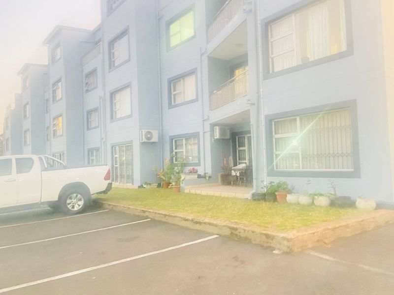 Exterior view of a multi-story apartment building featuring blue facade, balcony railings, security bars, and ground-level parking bays.