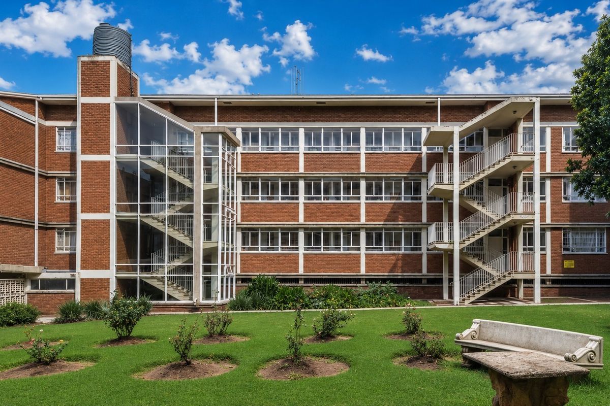 A multi-story commercial brick building featuring external staircases, large windows, and a well-maintained garden area with a stone bench.