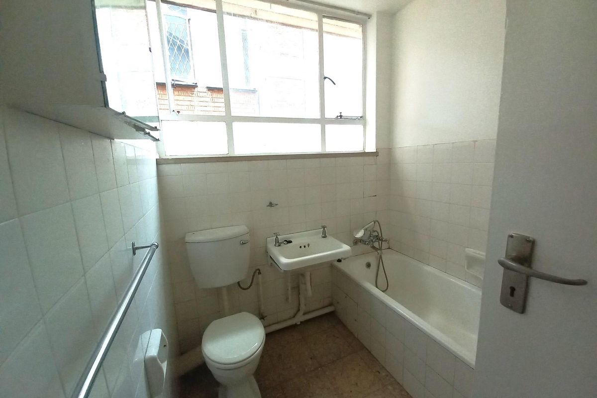 A compact, dated bathroom featuring a standard bathtub, wall-mounted basin, and toilet with white wall tiling and a large window for natural light.