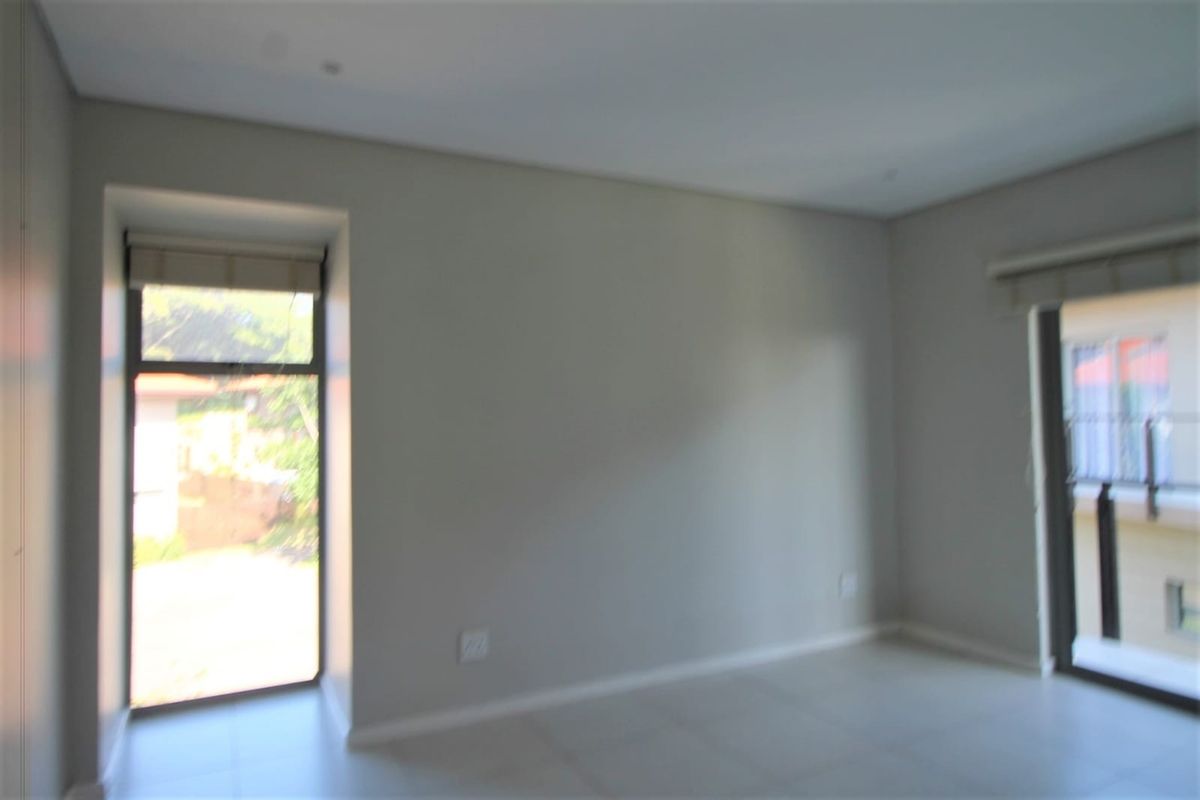 A bright, empty bedroom featuring neutral wall finishes, tiled flooring, and large windows providing natural light and access to an outdoor area.