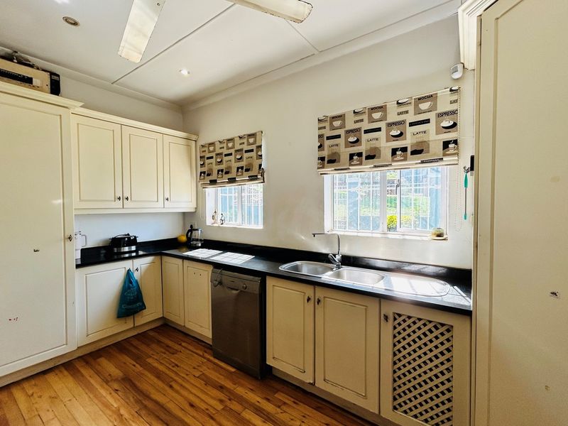 A functional kitchen featuring cream-colored cabinetry, dark countertops, a double stainless steel sink, and hardwood flooring with natural light from two windows.