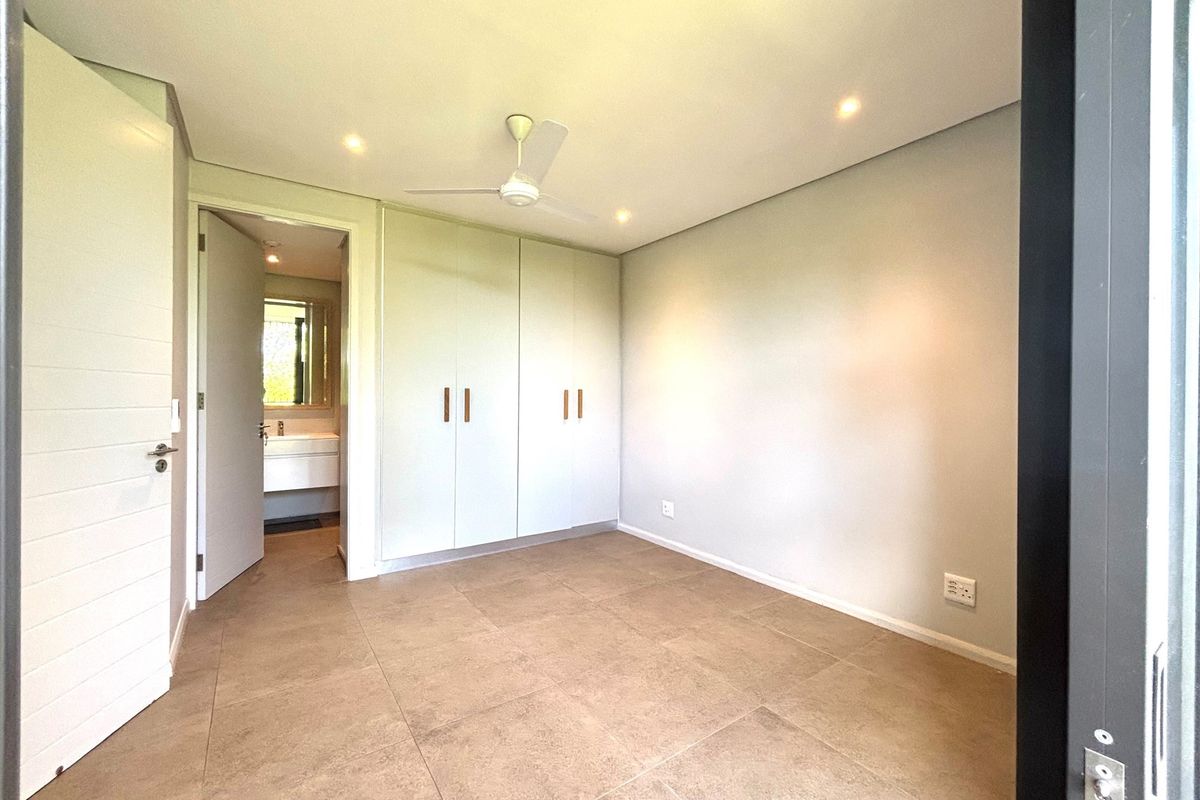 A bright, empty bedroom featuring neutral floor tiles, built-in cupboards, a ceiling fan, and an adjoining en-suite bathroom.
