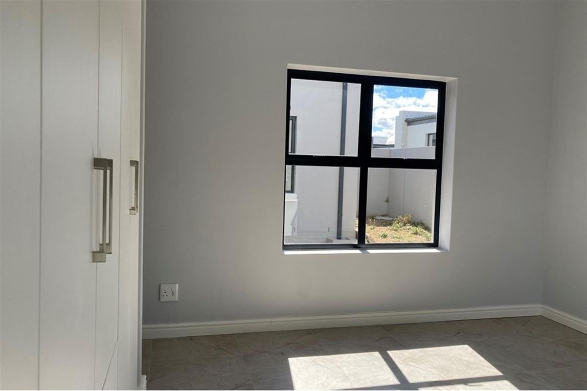 A bright, empty bedroom featuring modern tiled flooring, built-in cupboards, and a black aluminium window frame overlooking the exterior.