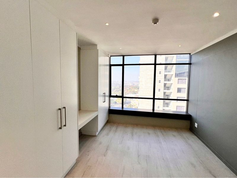 A bright, modern bedroom featuring built-in cupboards, a built-in desk, laminate flooring, and large windows offering expansive city views.