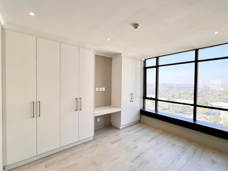 Modern bedroom featuring extensive built-in cabinetry, a dedicated integrated desk area, and expansive floor-to-ceiling windows offering panoramic city views.