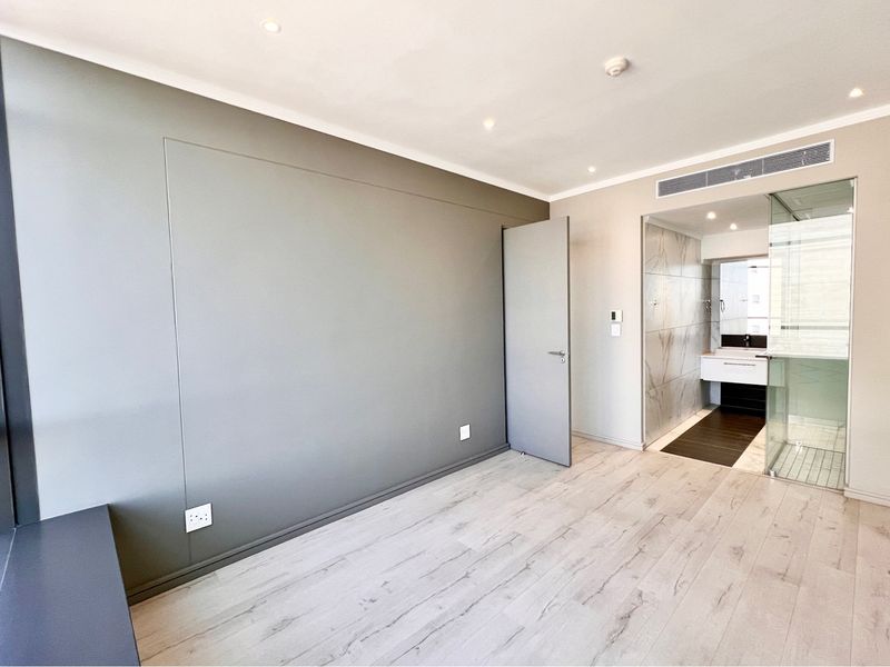 Modern bedroom featuring light laminate flooring, floor-to-ceiling windows, and an integrated en-suite bathroom with a glass shower enclosure.