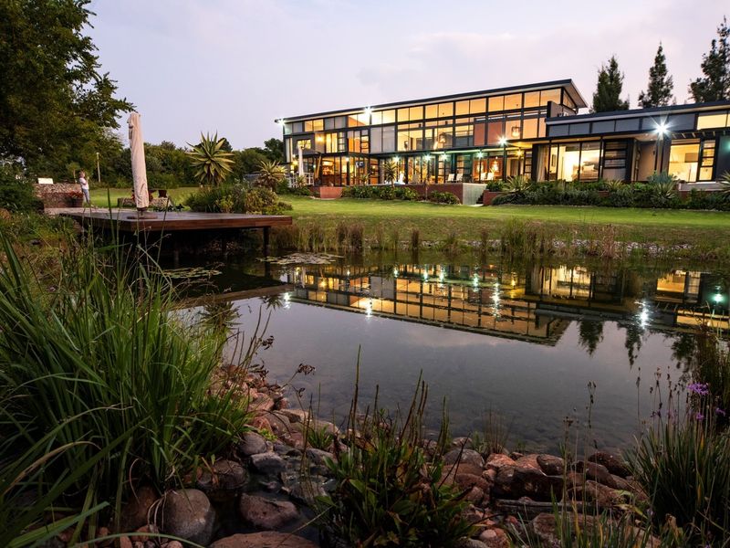 Stunning modern architectural home featuring an expansive glass facade overlooking a tranquil natural pond and beautifully landscaped garden.