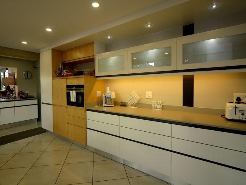 Modern kitchen featuring handleless white cabinetry, wood-grain accents, stone countertops, integrated oven, and ambient under-cabinet lighting with tiled flooring.