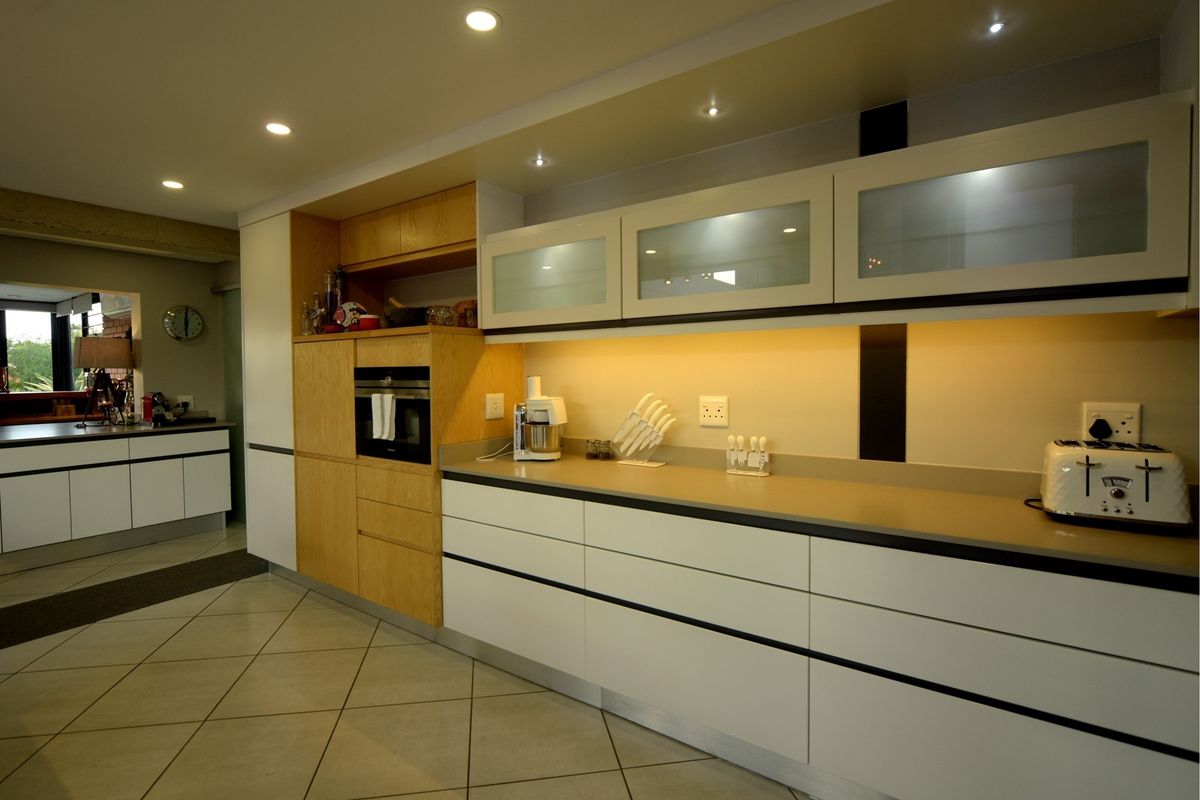 Modern kitchen featuring handleless white cabinetry, wood-grain accents, stone countertops, integrated oven, and ambient under-cabinet lighting with tiled flooring.