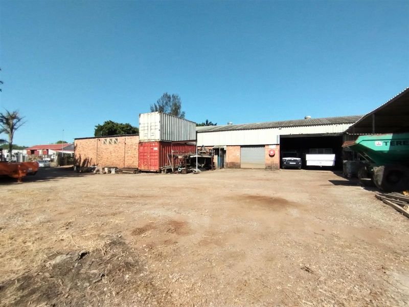 Industrial property exterior featuring a large unpaved yard area, brick warehouse structure, and roller shutter door access.