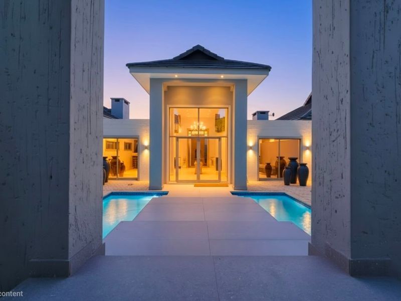 A stunning rear exterior view featuring a central swimming pool, paved walkway, and a grand glass entrance framed by modern architectural pillars.
