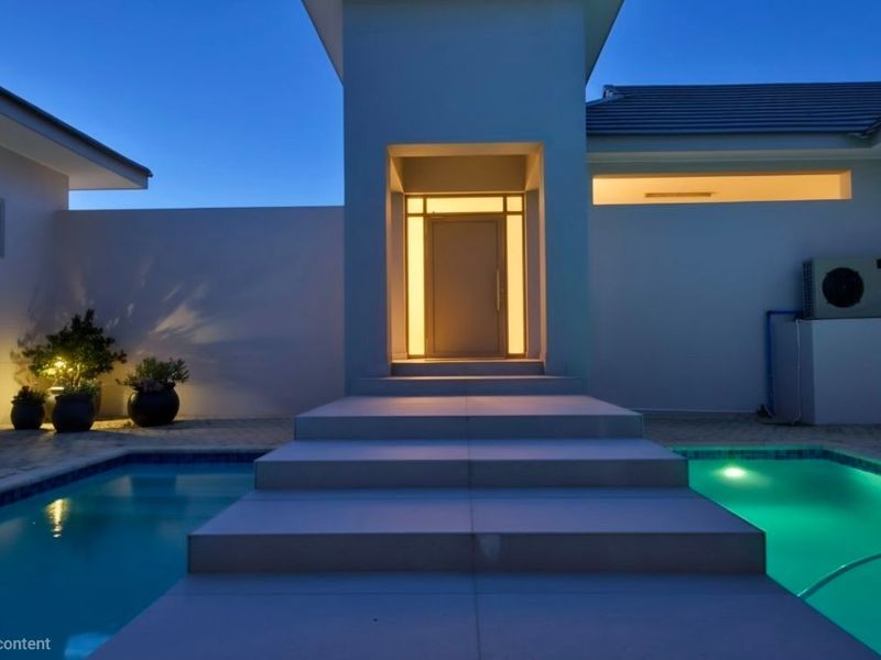 A modern exterior pool area at dusk, featuring a central stone walkway leading to an illuminated entrance and integrated pool lighting.