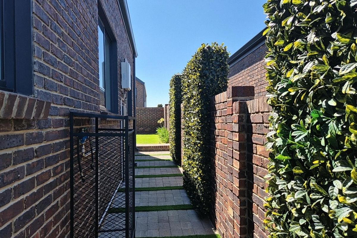A well-maintained side passage featuring brick walls, paved walkway with grass inserts, and manicured hedge screening.
