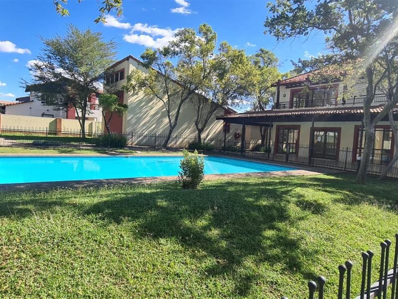 A well-maintained outdoor pool area featuring a clear swimming pool, surrounding lawn, and metal perimeter fencing, set against a residential building backdrop.