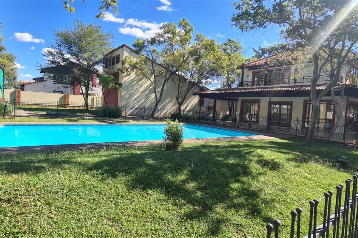 A well-maintained outdoor pool area featuring a clear swimming pool, surrounding lawn, and metal perimeter fencing, set against a residential building backdrop.