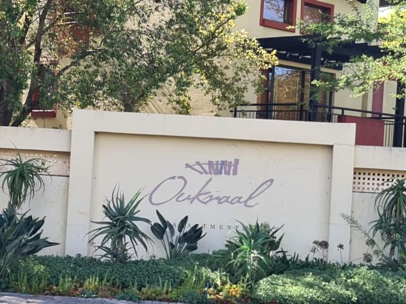 Front entrance view of the Oukraal apartment complex featuring a prominent boundary wall, signage, and established garden landscaping.