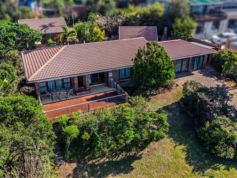 Aerial view of a single-story brick house featuring a tiled roof, a spacious wooden deck, and a well-established garden with a paved driveway.