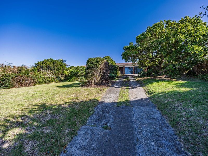 A front view of the property featuring a concrete driveway leading to the house, surrounded by a large, established garden with mature trees.