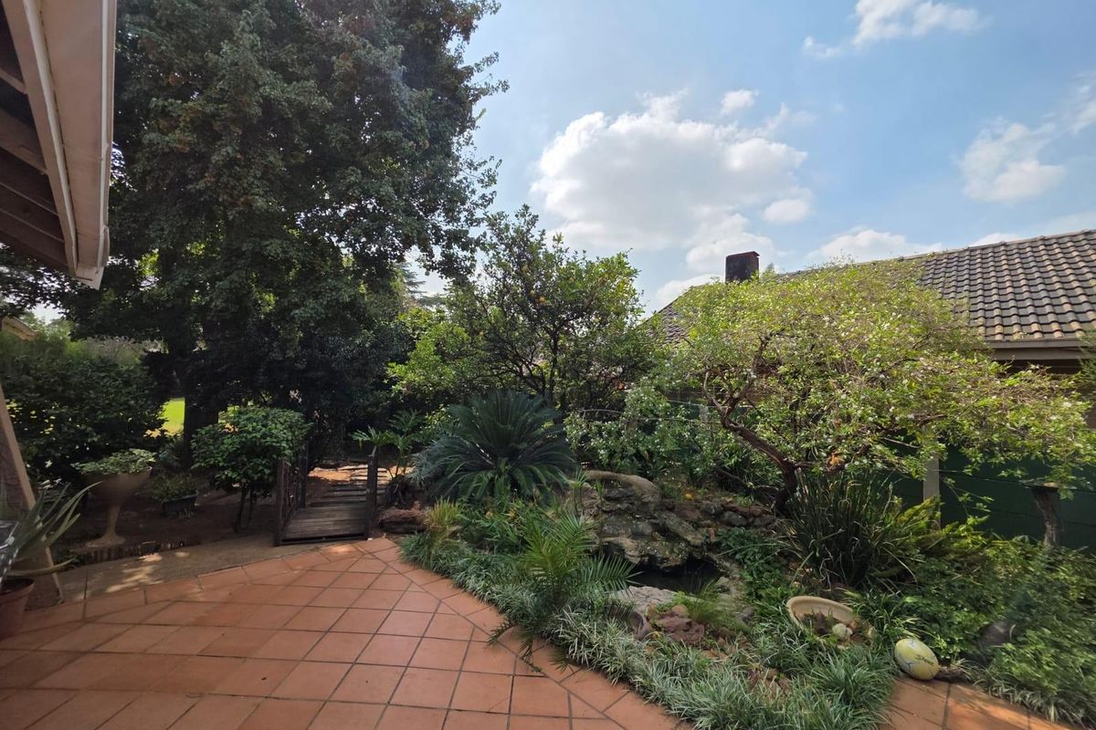 A landscaped rear garden featuring a tiled patio, a small decorative pond, and a wooden footbridge set within a mature, leafy outdoor space.
