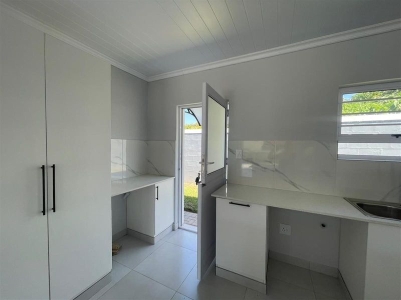 A clean, modern scullery or laundry area featuring white built-in cabinetry, stone countertops, a stainless steel sink, and direct access to the exterior.