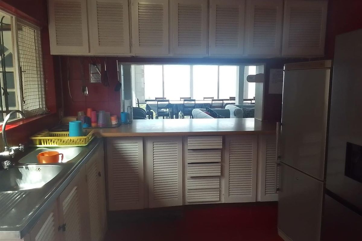 A dated kitchen featuring white louvered cabinetry, a stainless steel sink, and a serving hatch overlooking the adjacent living area.