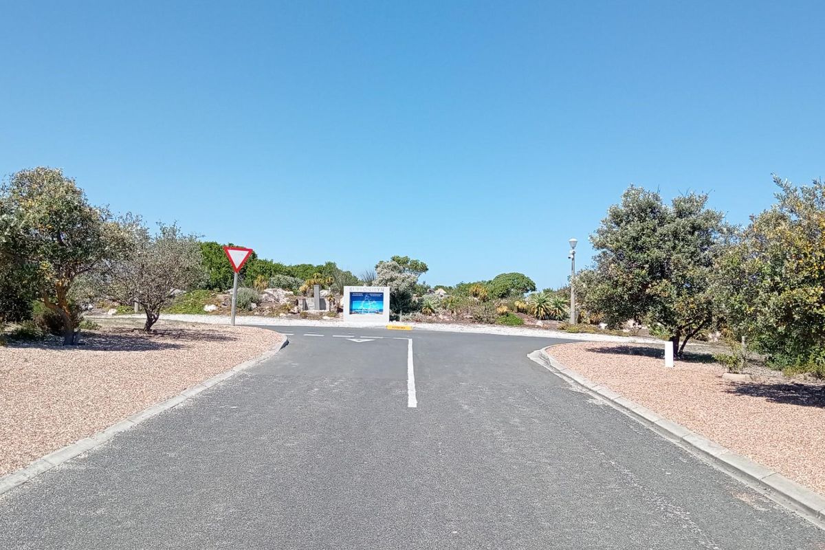 A clear street view showing the paved road, verge, and entrance signage for the residential estate.