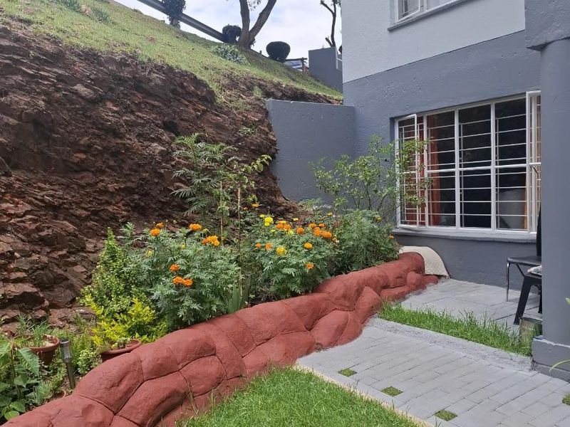 A private rear garden area featuring a paved walkway, a decorative stone-look retaining wall, and a landscaped lawn adjacent to the apartment building.