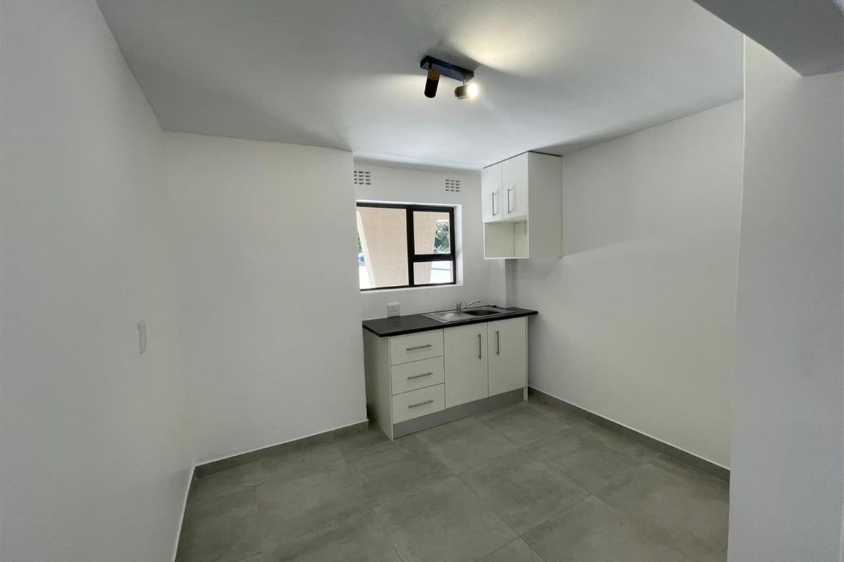 A clean, minimalist kitchen area featuring white built-in cabinetry, a dark countertop with a stainless steel sink, and modern grey floor tiles.