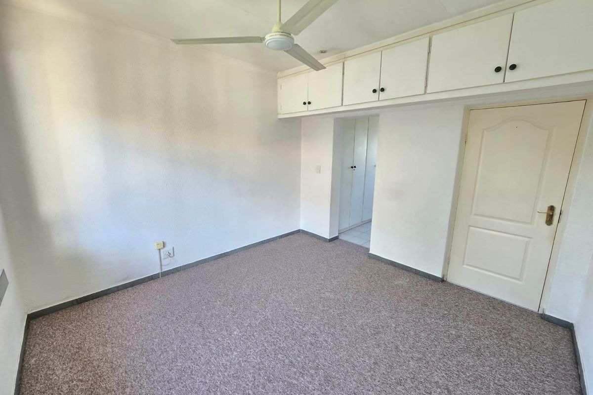A bright, empty bedroom featuring built-in overhead storage cupboards, a ceiling fan, and neutral carpet flooring.