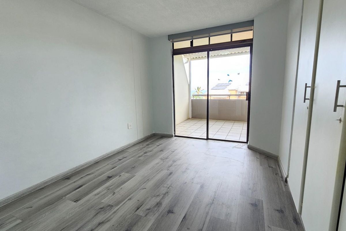 A bright, empty bedroom featuring modern laminate flooring, built-in cupboards, and a sliding glass door providing access to a private balcony.