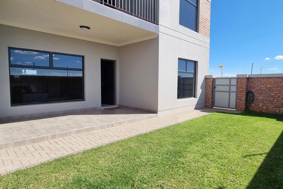A well-maintained rear exterior view featuring a paved patio area, a private lawn, and a brick boundary wall with security fencing.