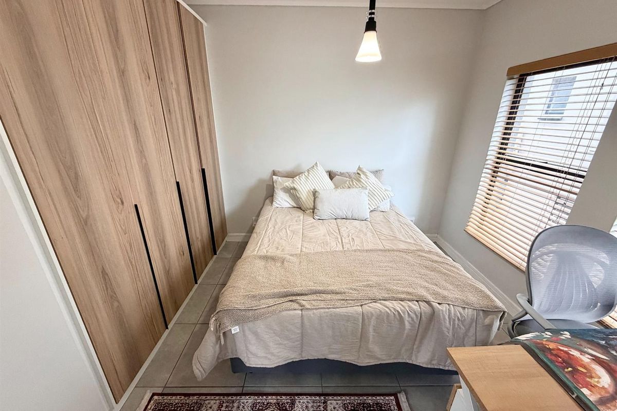 A well-maintained bedroom featuring modern wood-finish built-in cupboards, neutral tiled flooring, and natural light filtered through venetian blinds.