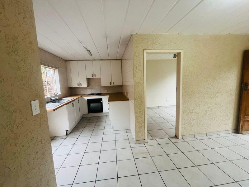 A dated kitchen featuring white cabinetry, built-in oven and hob, and tiled flooring, leading into an adjacent room.
