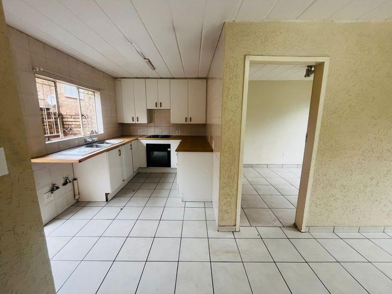 A dated kitchen featuring white cabinetry, built-in oven and hob, tiled flooring, and a double sink, adjacent to an open-plan living area.