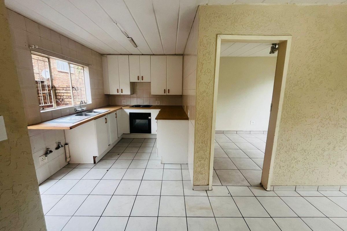 A dated kitchen featuring white cabinetry, built-in oven and hob, tiled flooring, and a double sink, adjacent to an open-plan living area.