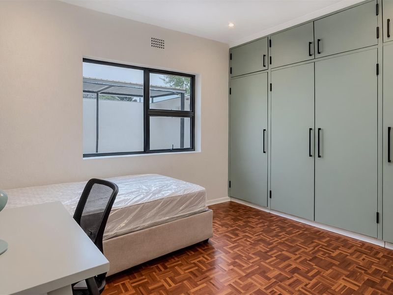 A bright bedroom featuring polished parquet flooring, built-in sage green cabinetry, and a large window providing natural light.