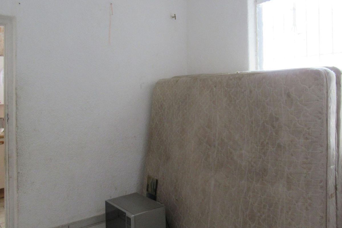A sparse bedroom featuring textured walls and a window with security bars, with a view into the adjacent kitchen area.