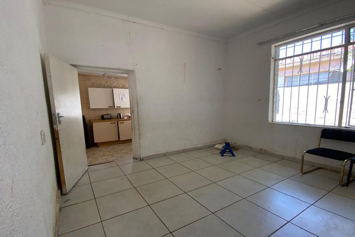 A sparsely furnished living area featuring tiled flooring and a window with security bars, leading into a separate kitchen space.