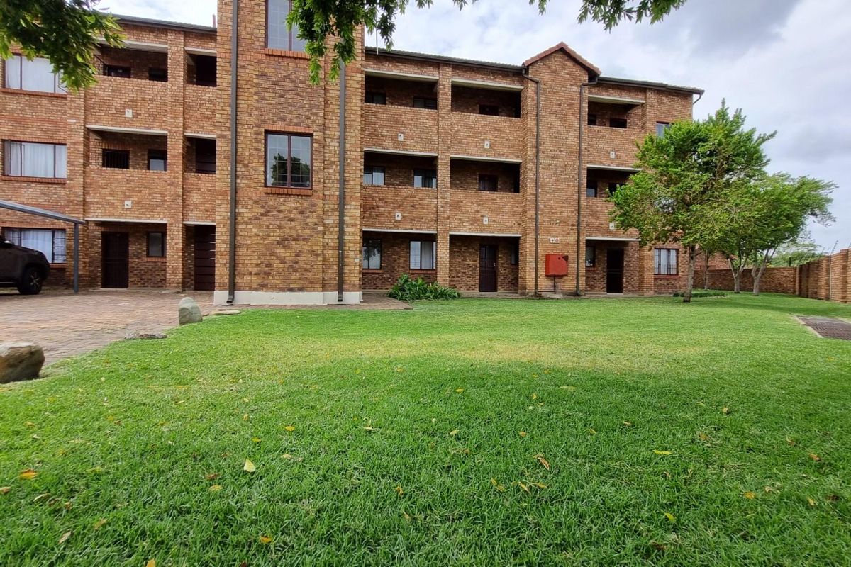 A view of the rear exterior of a multi-story face brick apartment complex featuring balconies, a paved driveway, and a well-maintained lawn.