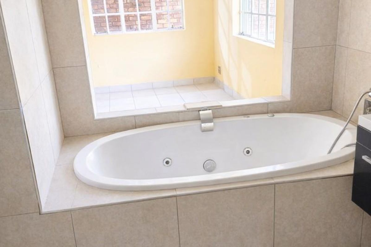 A bathroom featuring a built-in jetted bathtub set within a tiled surround, positioned against a large window overlooking an adjacent sunroom.