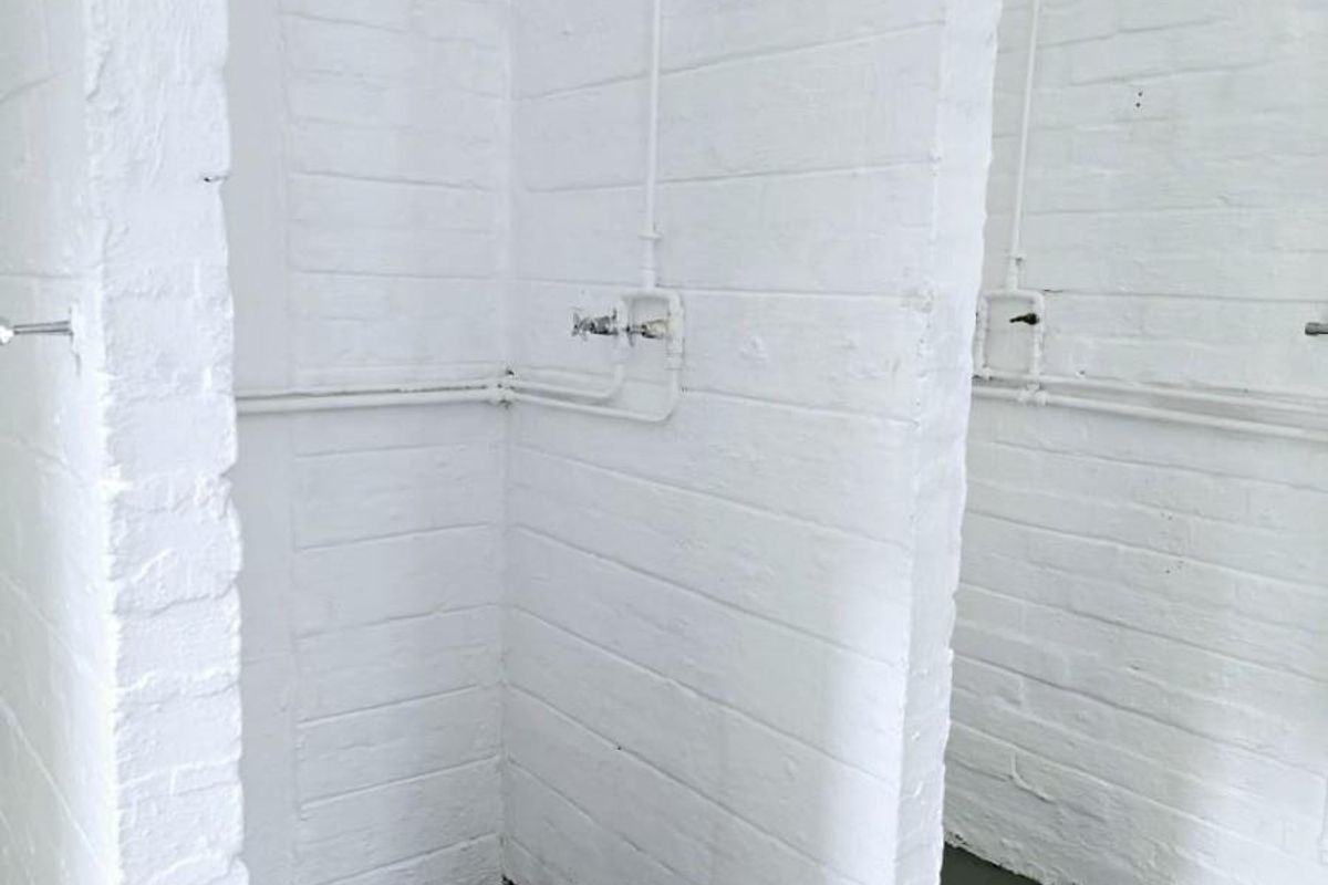 Industrial-style communal shower area featuring exposed white-painted brick partitions, wall-mounted shower heads, and concrete flooring.