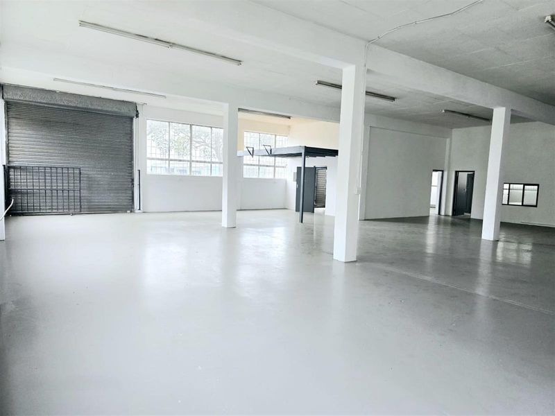 Spacious open-plan industrial warehouse featuring concrete flooring, high ceilings with fluorescent lighting, a roller shutter door, and an integrated mezzanine level.