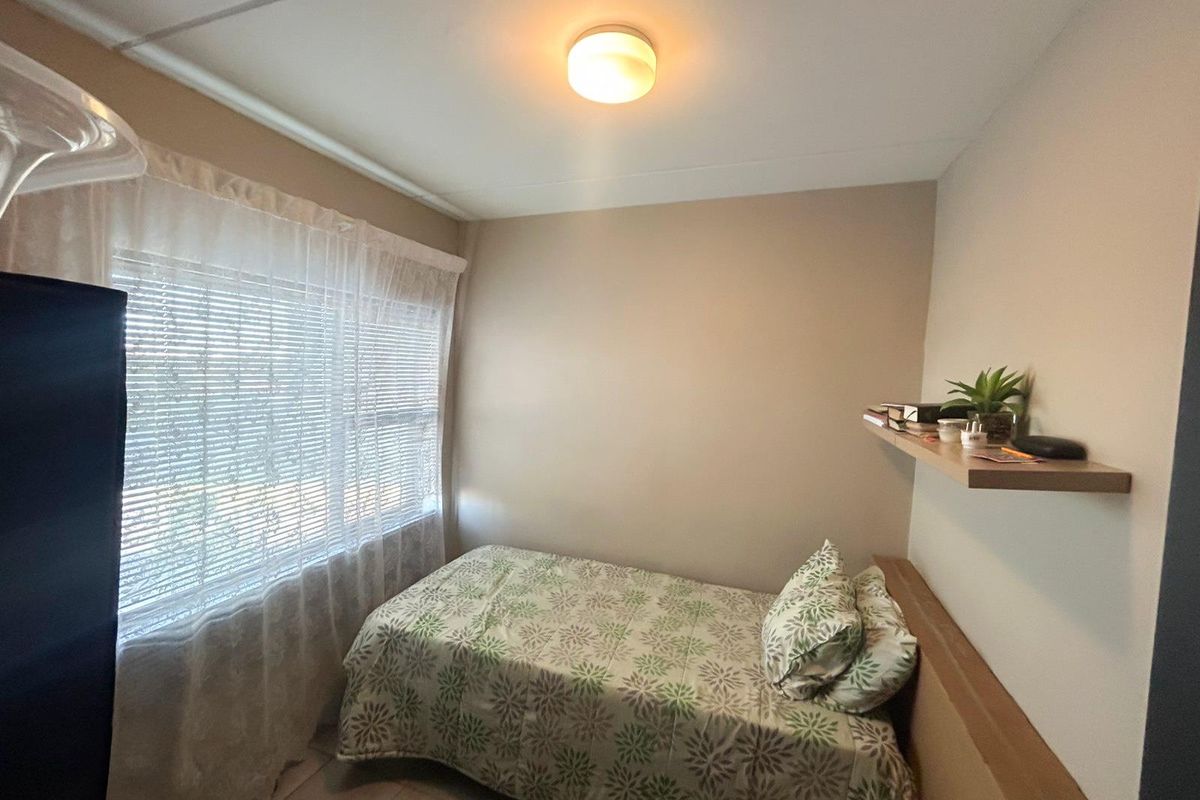 A compact bedroom featuring a window with blinds, a ceiling-mounted light fixture, and a wall-mounted floating shelf.