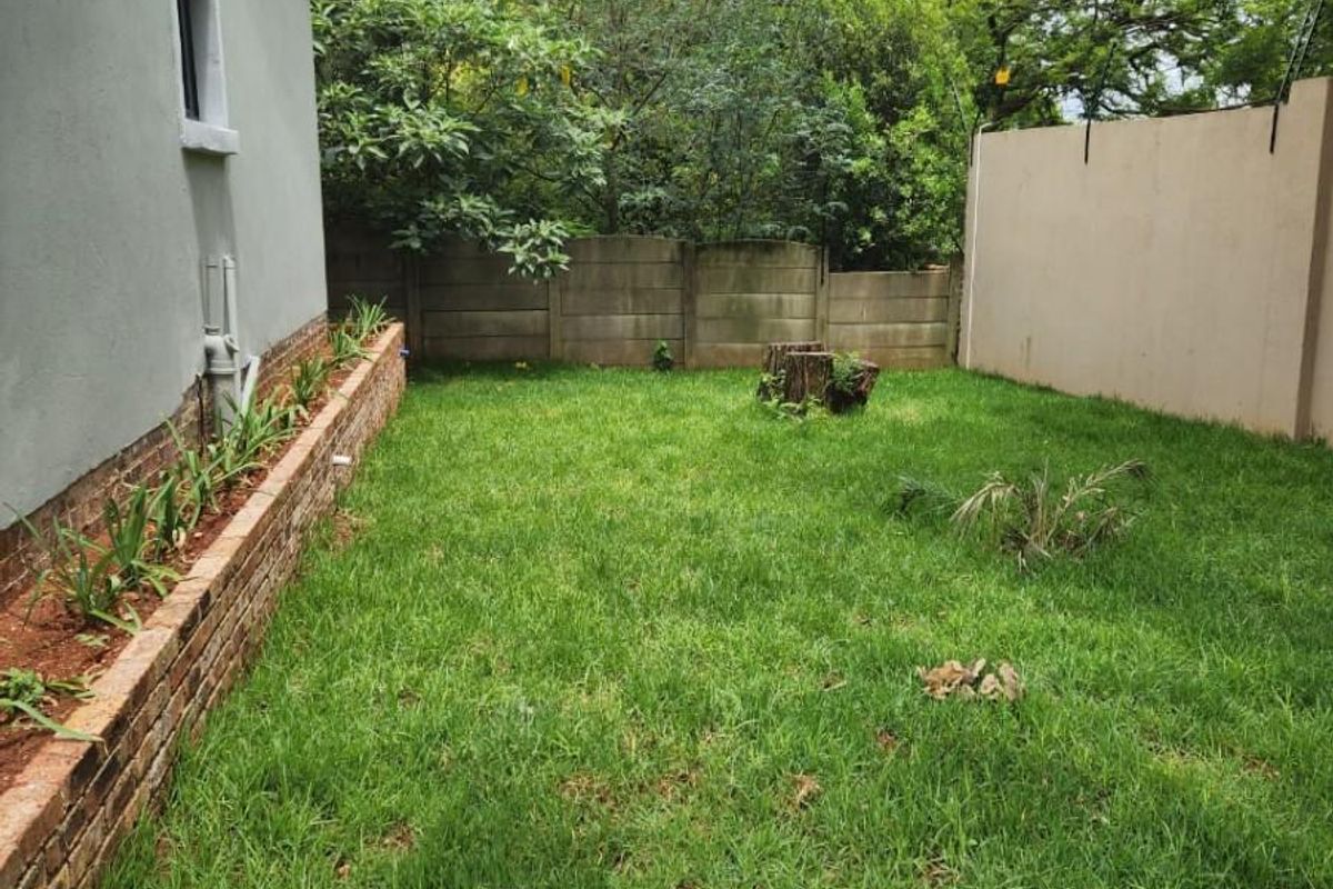 A private rear garden featuring a brick-edged planter bed, a grass lawn, and perimeter boundary walls with mature trees in the background.