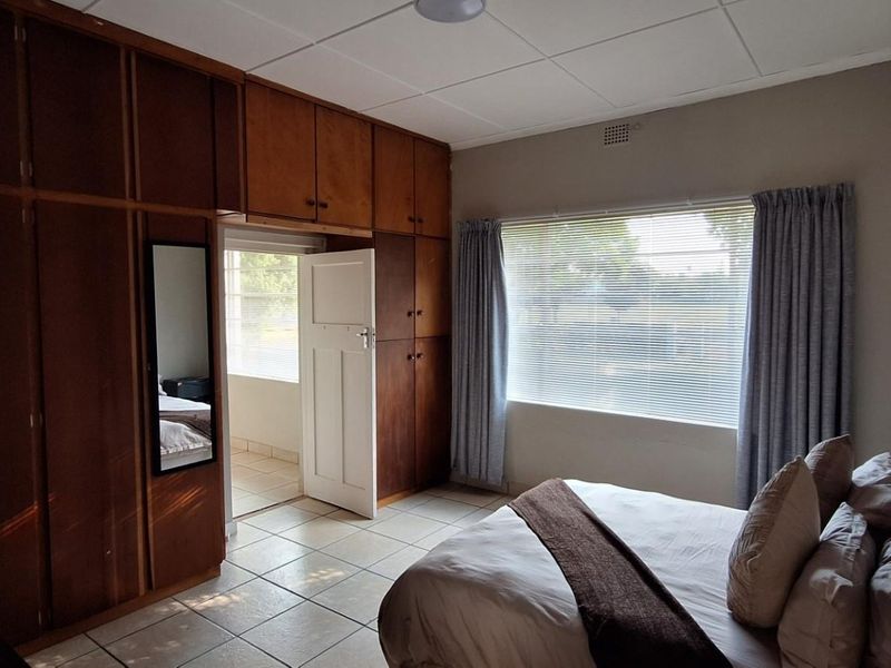 A bedroom featuring extensive floor-to-ceiling built-in wooden cupboards, tiled flooring, and a large window with blinds.