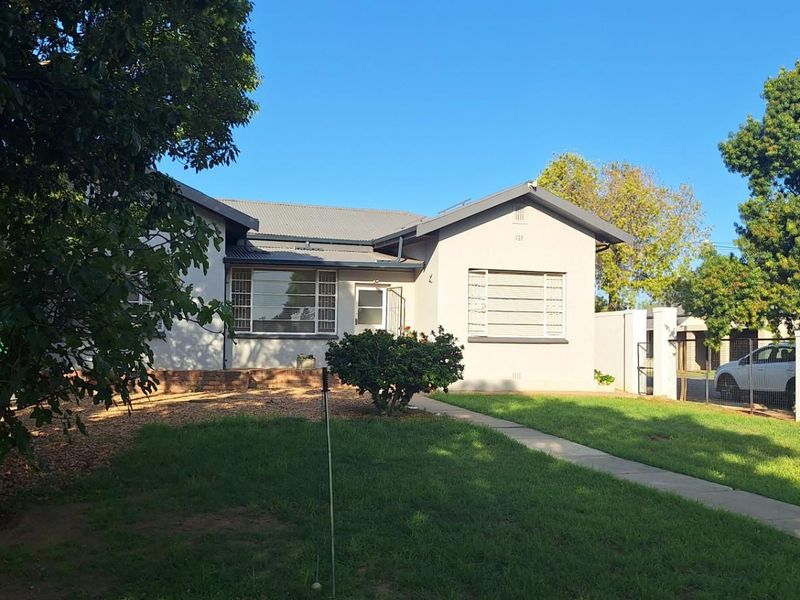A single-story residential house featuring a front garden, paved walkway, and metal-framed windows, set within a fenced property boundary.
