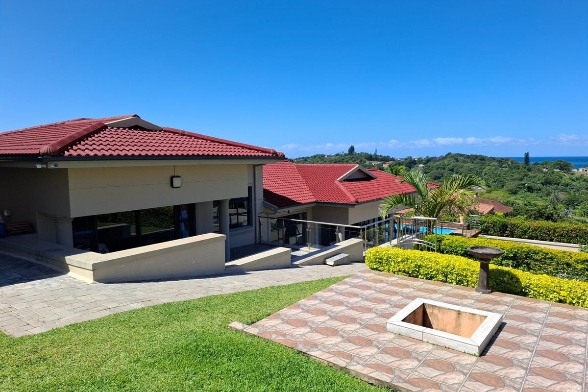 A spacious rear exterior view featuring a paved patio, landscaped garden, swimming pool, and scenic coastal views under a clear blue sky.