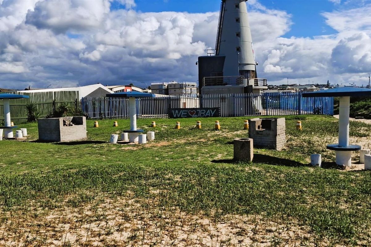 Outdoor recreational area featuring fixed concrete braai stands and shaded picnic tables, situated near a lighthouse and coastal fencing.