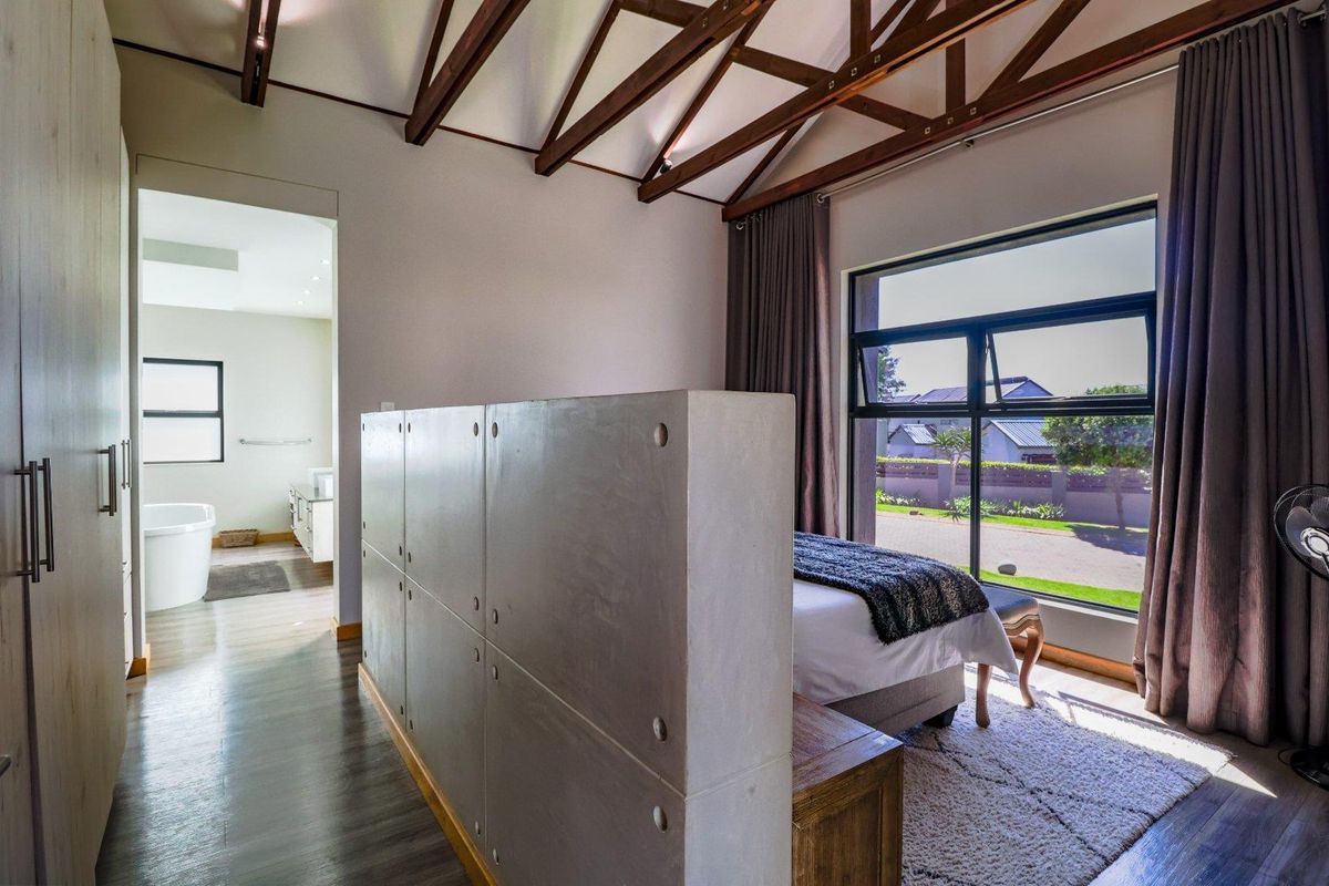 Spacious bedroom featuring exposed timber roof trusses, a modern concrete partition wall, built-in wardrobes, and large windows with views of the surrounding area.
